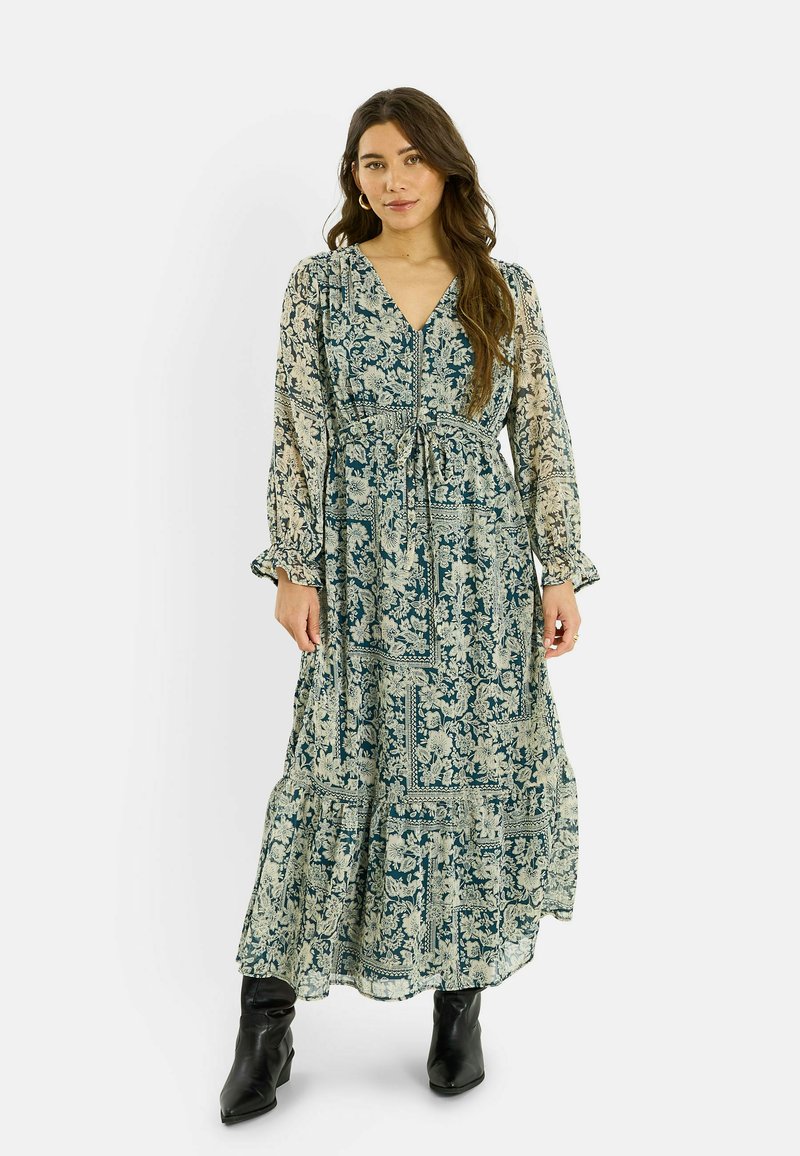 Robe fluide longue en imprimé floral bleu et crème, avec un décolleté en V, une taille cintrée, des manches longues et un ourlet à volants, associée à des bottes noires.