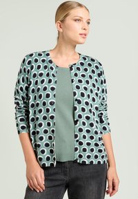 Kobieta z blond włosami nosi zielony kardigan w czarno-białe kropki na jednolitym zielonym topie, połączony z czarnymi dżinsami, na jednolitym tle.