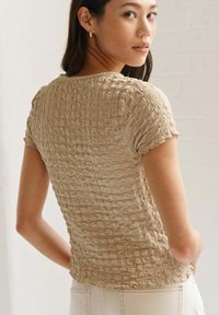 Haut texturé beige à manches courtes, présentant un design froncé et un col rond, porté avec un pantalon clair à taille haute.