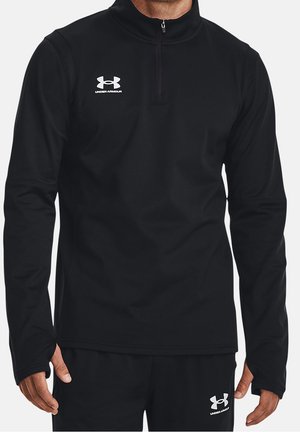 Hombre con camiseta negra de manga larga Under Armour con cremallera hasta el pecho y pantalones negros a juego con logotipos blancos de Under Armour en el pecho y el muslo.