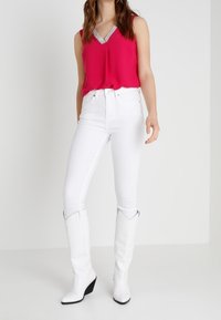 Fuchsia ärmlös topp med en paljetterad V-ringning, kombinerad med figurnära vita jeans och vita kilklackar.