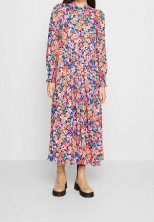 Robe florale avec un fond bleu vif et des motifs de fleurs roses et oranges, manches longues et col haut. Chaussures à botte montrées.