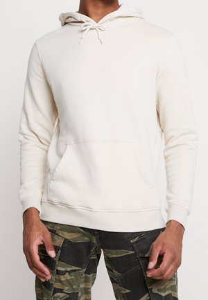 Mand iført en off-white hættetrøje med forlommer og camouflage cargobukser, stående foran en ensfarvet baggrund.
