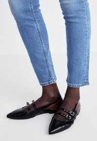Schwarze Lederflats mit spitzen Zehen, zwei Riemen mit silberner Hardware und einem niedrigen Absatz, kombiniert mit eng anliegenden blauen Jeans.