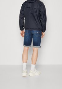 Chaqueta cortavientos azul marino sobre pantalones cortos de mezclilla azul con dobladillos enrollados. Calcetines blancos y zapatillas de color claro completan el conjunto.