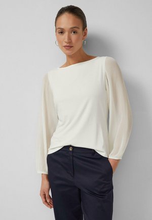 Frau trägt eine weiße Bluse mit durchsichtigen, plissierten langen Ärmeln und marineblaue Hose, steht mit einer Hand in der Tasche vor einer schlichten Wand.