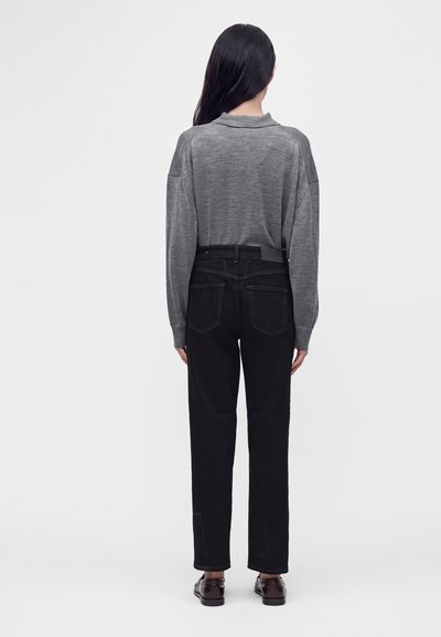 Personne aux longs cheveux foncés portant un pull gris à manches longues, un jean noir taille haute et des mocassins foncés, debout de dos.