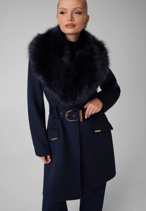 Femme portant un manteau bleu marine avec un grand col en fourrure noire, ceinture à boucle dorée et bijoux en or, posant devant un fond gris uni.