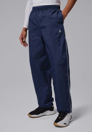 Hombre vistiendo pantalones deportivos azul marino con ribete blanco y logo de Jordan, combinado con zapatillas negras y blancas, de pie con una mano en el bolsillo.