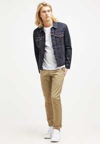 Donkerdenim jacket met twee borstzakken, gecombineerd met een wit t-shirt en beige chinos. Witte sneakers maken de outfit compleet.
