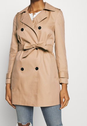 Trench-coat beige croisé avec des boutons noirs et une ceinture nouée à la taille, porté sur un haut blanc et un jean bleu déchiré.