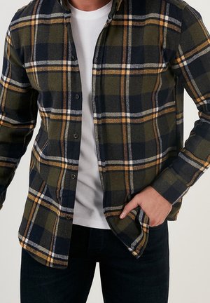 Man met een groen, zwart en geel geruit overhemd over een wit T-shirt, met één hand in zijn zak en zwarte broek.