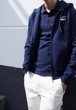 Homme portant un polo Lacoste bleu marine et un sweat à capuche zippé avec un pantalon blanc, debout devant un mur blanc et noir.