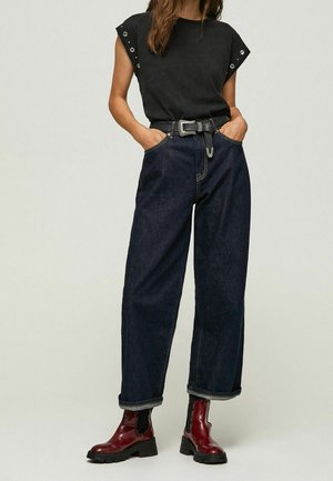 Kobieta ubrana w czarny bezrękawnik ze szlufkami, ciemnoniebieskie dżinsy z wysokim stanem i czarnym paskiem oraz błyszczące ciemnoczerwone botki za kostkę.