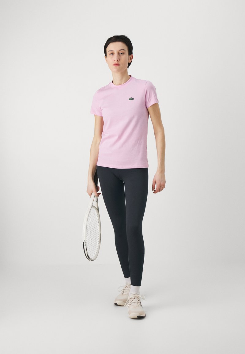 T-shirt in cotone rosa chiaro abbinata a leggings neri, con in mano una racchetta da tennis bianca. Presenta un logo verde sulla maglietta.