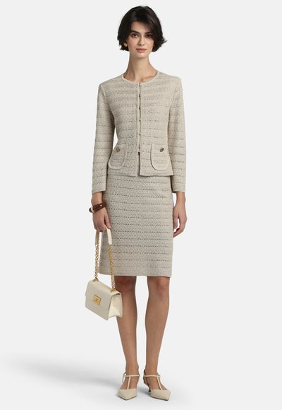 Femeie purtând o jachetă bej texturată cu nasturi și o fustă asortată până la genunchi, ținând o geantă crem cu lanț auriu, stând pe un fundal alb.