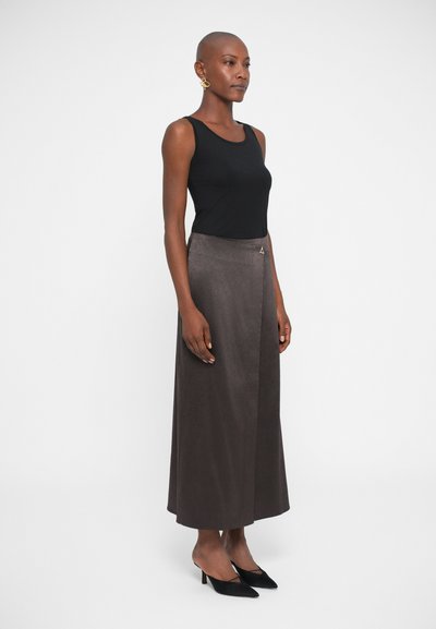 Mujer vestida con una blusa sin mangas negra, una falda larga marrón oscuro y tacones negros de punta, de pie frente a un fondo blanco liso.