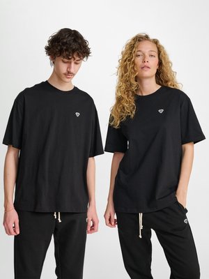 Man en vrouw dragen bijpassende zwarte oversized t-shirts en zwarte broeken met kleine witte logo's, staand tegen een effen witte achtergrond.