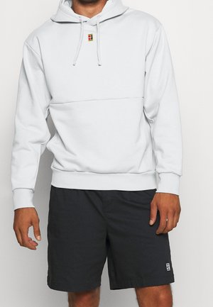 Persona che indossa una felpa con cappuccio bianca con coulisse e un piccolo logo, abbinata a pantaloncini neri con una piccola toppa sulla gamba sinistra.