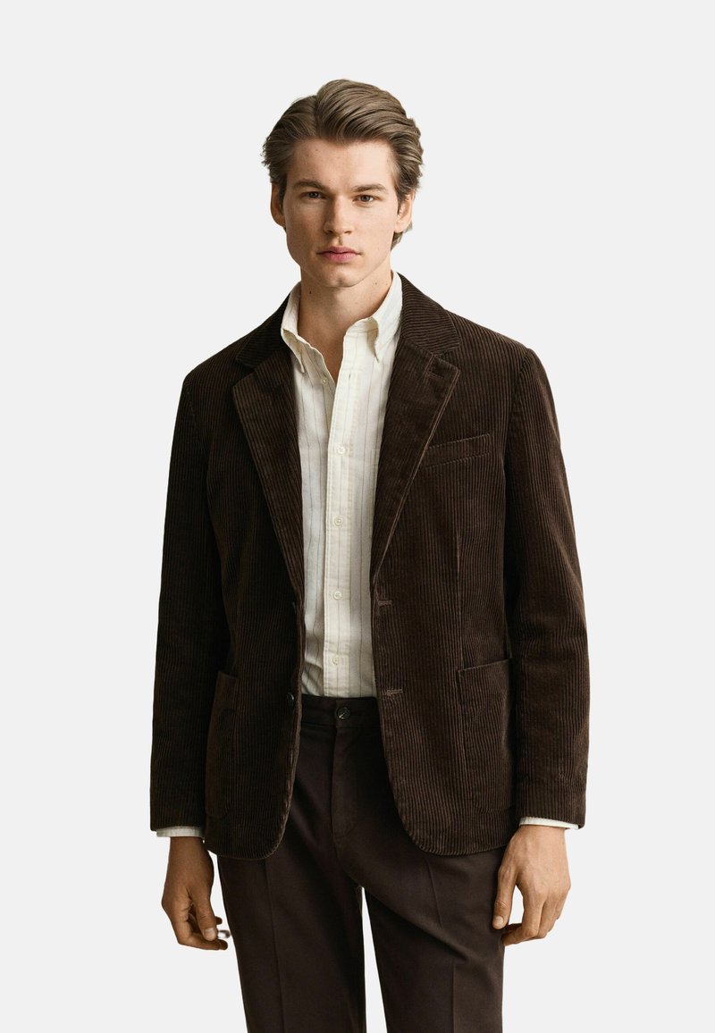 Jeune homme aux cheveux bruns coiffés porte un blazer en velours côtelé marron foncé, une chemise blanche boutonnée et un pantalon foncé sur un fond clair.