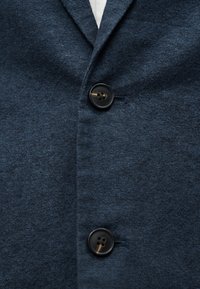 Σκούρο μπλε σακάκι με υφή, με δύο μαύρα κουμπιά κουμπωμένα πάνω από γιακά λευκού πουκάμισου που φαίνεται στην κορυφή.