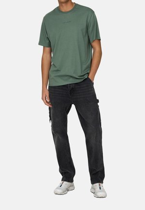 Grünes Baumwoll-T-Shirt mit Rundhalsausschnitt, kombiniert mit schwarzen Jeans aus Denim mit Seitentaschen. Leichte Sneaker runden das Outfit ab.