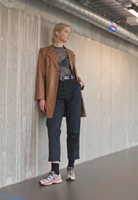 Brown patterned oversized blazer, black fitted top, black tapered pants, and multi-colored sneakers. Gray background with concrete wall.