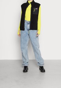 Black fleece vest with a zip front and a chest pocket, paired with a bright yellow long-sleeve top and light blue baggy jeans; black boots.
