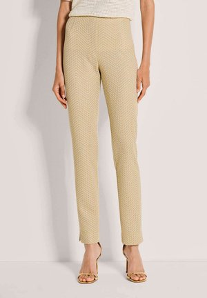 Beige strukturierte, schmal geschnittene Hose getragen mit goldfarbenen Sandalen mit hohem Absatz, kombiniert mit einem cremefarbenen Strickoberteil, stehend vor einem schlichten hellen Hintergrund.