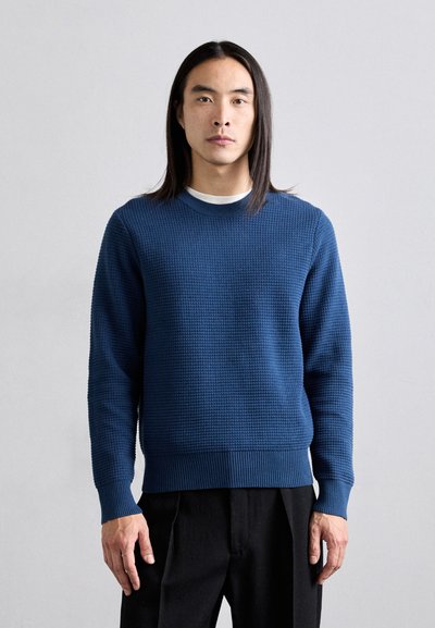 Pull bleu tricoté avec un motif texturé, encolure ronde et poignets et ourlet côtelés, porté sur un pantalon noir.