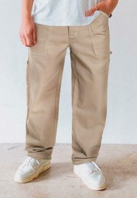 Beige Cargo-Hosen mit weißen Nähten, ausgestattet mit Seitentaschen und einem lockeren Schnitt, kombiniert mit weißen Sneakern auf einem strukturierten Boden.