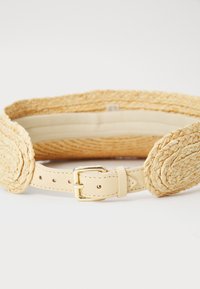 Flätad halmbälte i ljusbeige med en slät läderbaksida och ett metallic guldspänne. Har flera hål för storleksjustering.