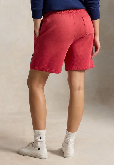 Personne portant un short rouge décontracté, des chaussettes blanches avec un logo et des baskets blanches, debout avec une main dans la poche devant un fond uni.