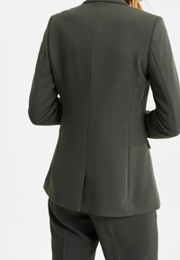 Tmavě zelené sako na míru s dlouhými rukávy, s jediným zadním rozparkem a elegantními liniemi. Vyrobeno z hladké, strukturované látky, s klasickým designem.