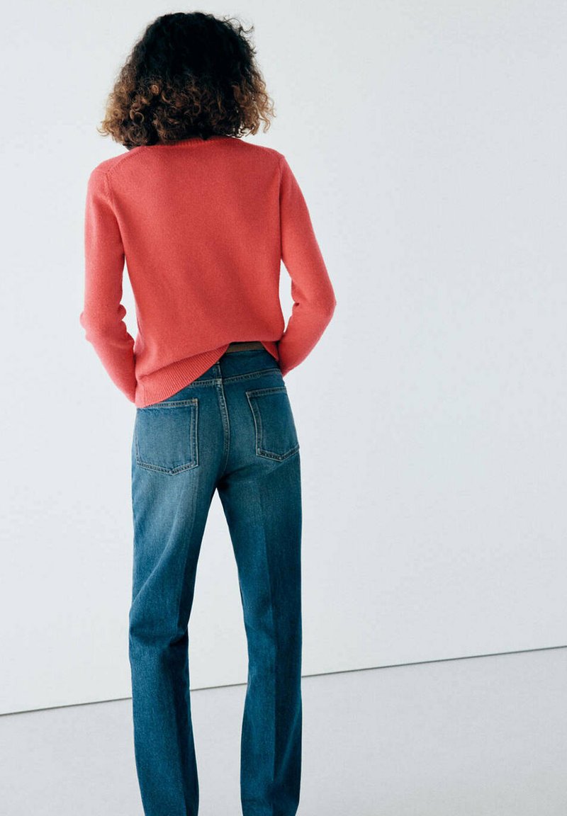 Personne aux cheveux bouclés vue de dos portant un pull corail et un jean bleu sur un fond clair uni.