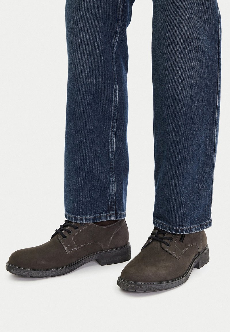Hnědé svrškové šněrovací boty s texturovaným povrchem, vybavené černou gumovou podrážkou a kontrastním prošíváním, spárované s tmavě modrými džíny.