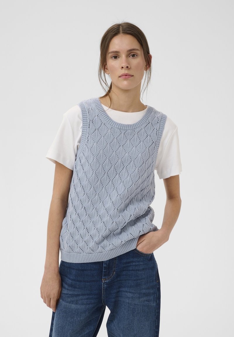 Femme portant un pull sans manches en tricot bleu clair par-dessus un t-shirt blanc et un jean bleu, debout avec une main dans sa poche.