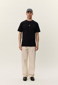 Melna īsas piedurknes T-krekls ar centrētu baltu "II" grafiku, kombinēts ar bēšām plašām biksēm un melniem apaviem, pabeigts ar pelēku cepuri.