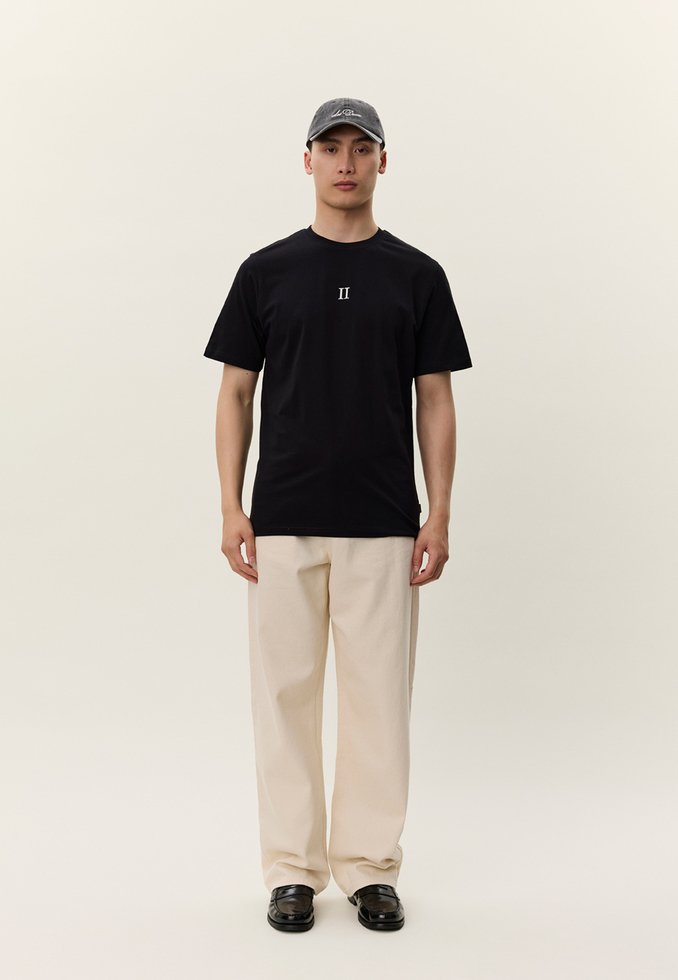 Melna īsas piedurknes T-krekls ar centrētu baltu "II" grafiku, kombinēts ar bēšām plašām biksēm un melniem apaviem, pabeigts ar pelēku cepuri.