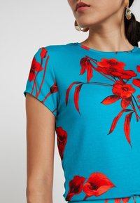 Top corto de color turquesa de manga corta, hecho de una tela suave, con un audaz estampado floral rojo y un cuello redondo, con una textura suave.