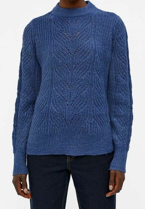 Pull en tricot torsadé bleu à manches longues porté avec un pantalon foncé, montrant des motifs tricotés texturés ainsi qu'un ourlet et des poignets côtelés.