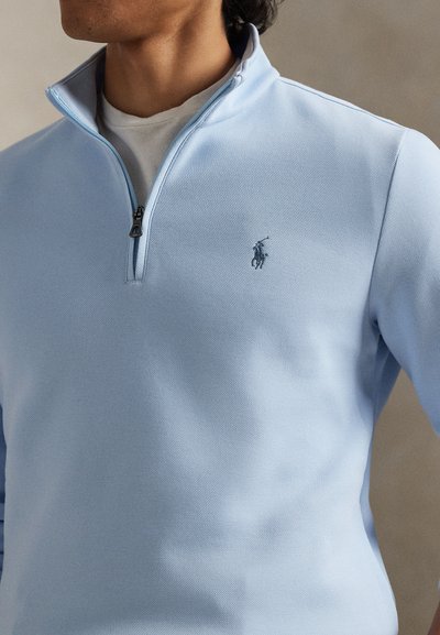Άνδρας που φοράει μπλε ανοιχτό πουλόβερ με φερμουάρ τεταρτημόριο πάνω από λευκή μπλούζα, με μικρό σκούρο κεντητό λογότυπο παίκτη πόλο στο στήθος.