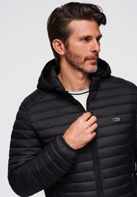 Chaqueta de plumas negra con capucha, con cierre de cremallera en la parte delantera, patrón acolchado horizontal y un pequeño logo en el pecho.
