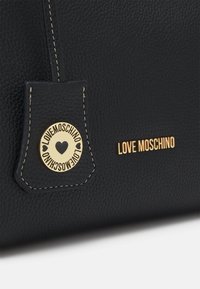 Svart, texturerad läderhandväska med guld "LOVE MOSCHINO"-bokstäver och rund guldemblem med hjärtan och varumärkesnamn.