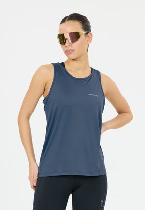 Kvinde iført en marineblå ærmeløs atletisk top og sorte leggings, poserer med den ene hånd på hoften og reflekterende store sports-solbriller.
