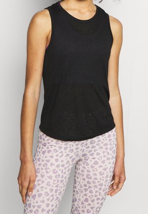 Vrouw met een mouwloos zwart topje en witte leggings met een lichtpaarse luipaardprint staand tegen een effen achtergrond.