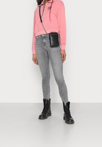 Felpa rosa con zip, jeans skinny grigi e stivali neri stringati. Pochette nera per il telefono a tracolla attaccata con una catena.