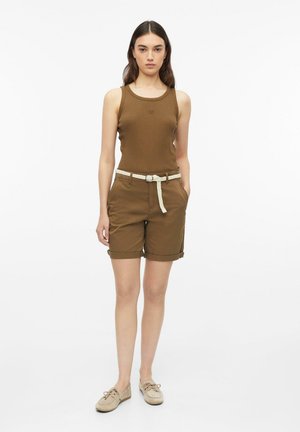 Vrouw die een bruine mouwloze top draagt, bijpassende shorts met een witte gevlochten riem en beige veterschoenen, staande tegen een witte achtergrond.