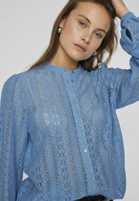 Blouse en dentelle bleu clair avec un devant à bouton, un col rond et des manches bouffantes. Présente des motifs floraux élaborés et une texture transparente.