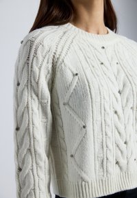 Nainen, joka käyttää valkoista neulottua villapaitaa, jossa on köysimäisiä kuvioita ja pieniä hopeisia helmiä etuosassa ja hihoissa.